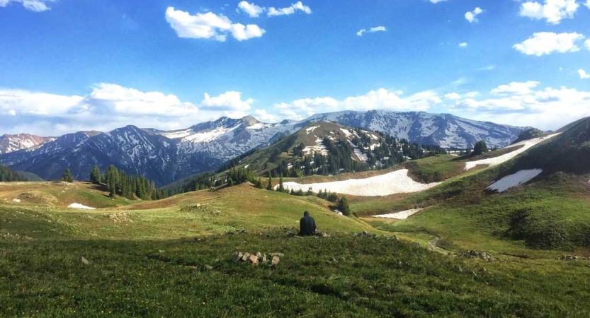 a person sits in a green alpine meadow, facing a snow-covered mountainous landscape.